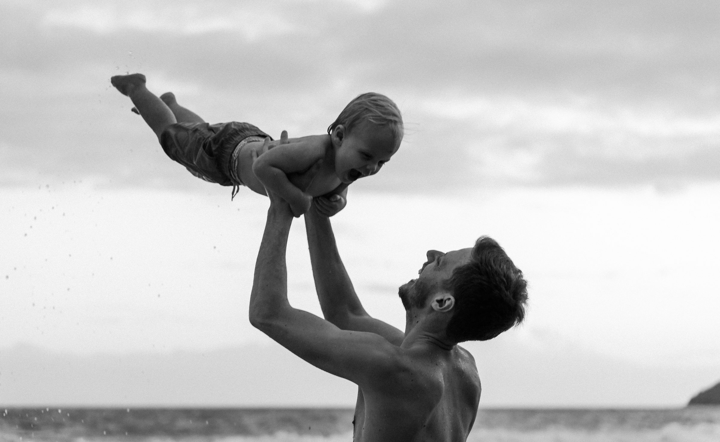 Enfant soulevé à bout de bras au bord de la mer, en noir et blanc